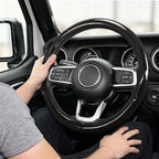 Installed view of glossy black carbon-fiber-style 3-piece steering wheel cover for Jeep Wrangler 2018-2026 with driver holding the wheel#color=glossy black